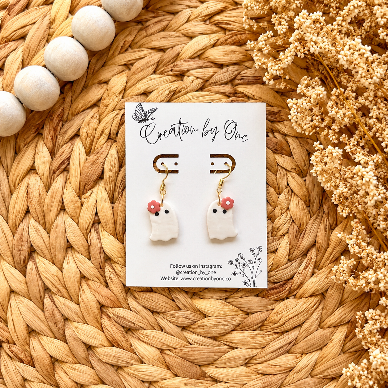 Pair of ghost-shaped earrings on a card labeled 'Creation by One' on a woven surface with dried flowers.