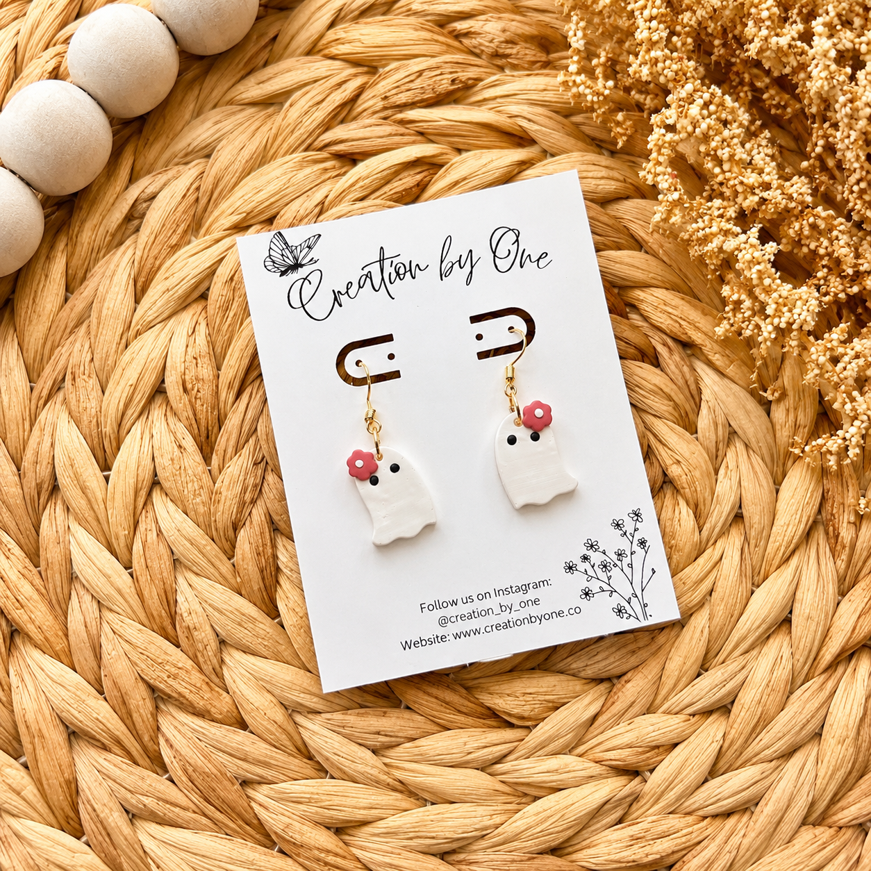 Pair of ghost-shaped earrings on a 'Creation by One' branded card, placed on a woven surface with dried flowers.