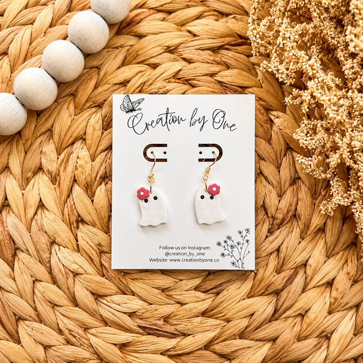 Pair of ghost-shaped earrings on a 'Creation by One' branded card, placed on a woven surface with decorative elements.