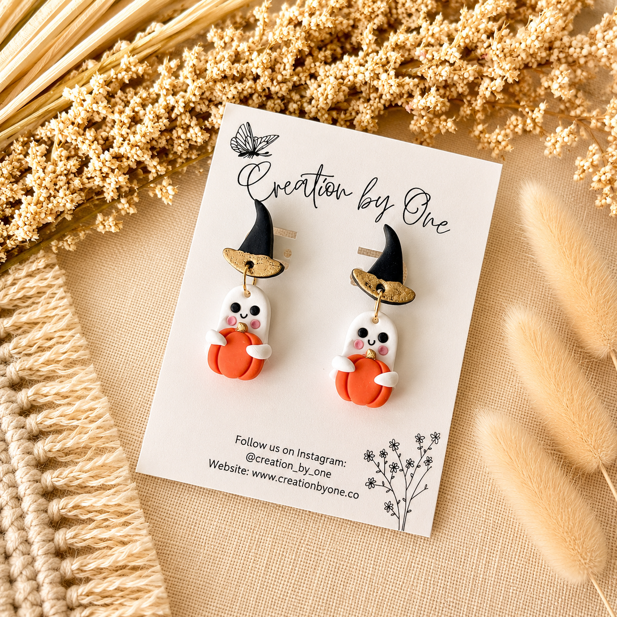 Halloween-themed earrings on a 'Creation by One' branded card with dried plants in the background.
