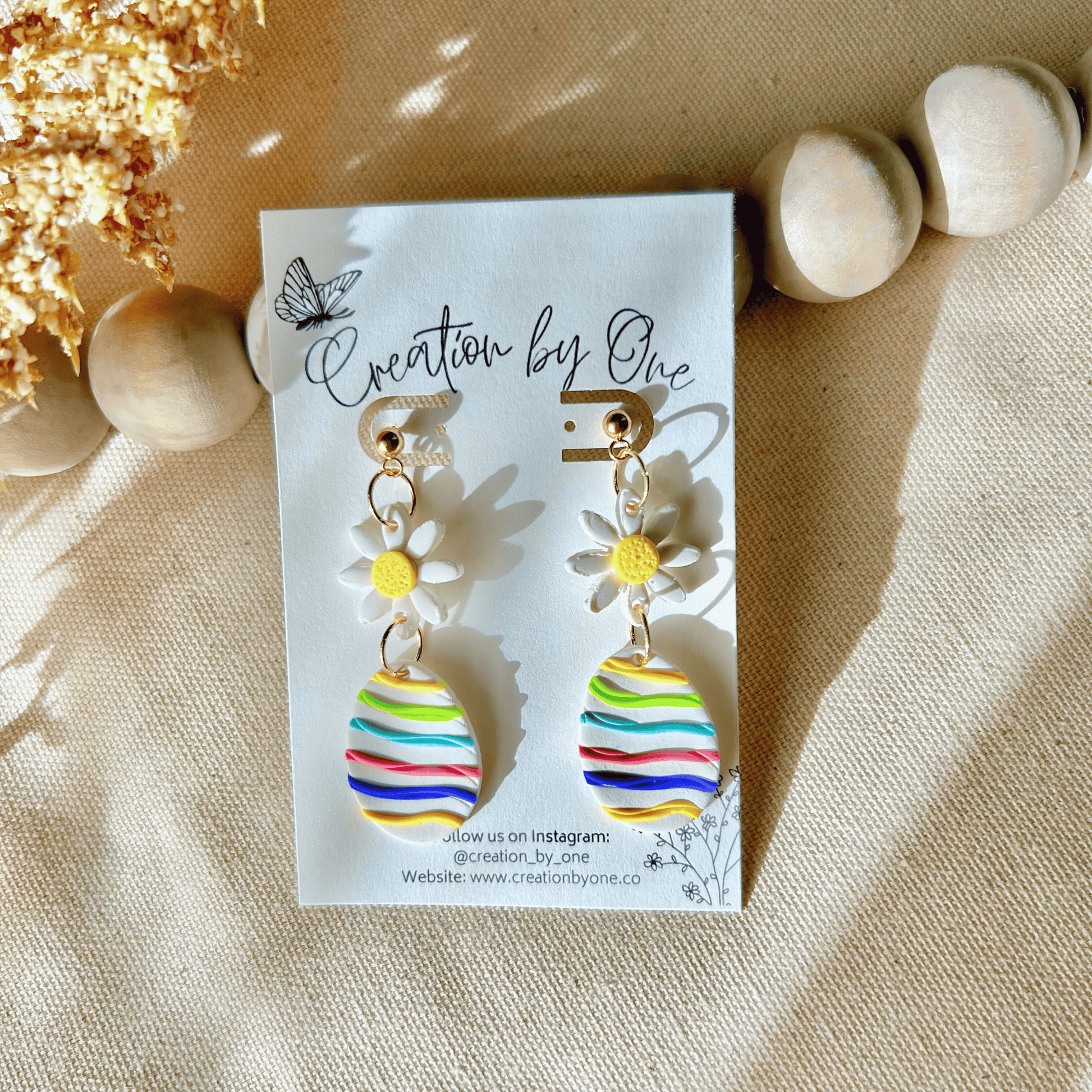 A 14K gold-plated, striped colorful egg-shaped earring with a daisy design at the top, displayed on a beige fabric surface.