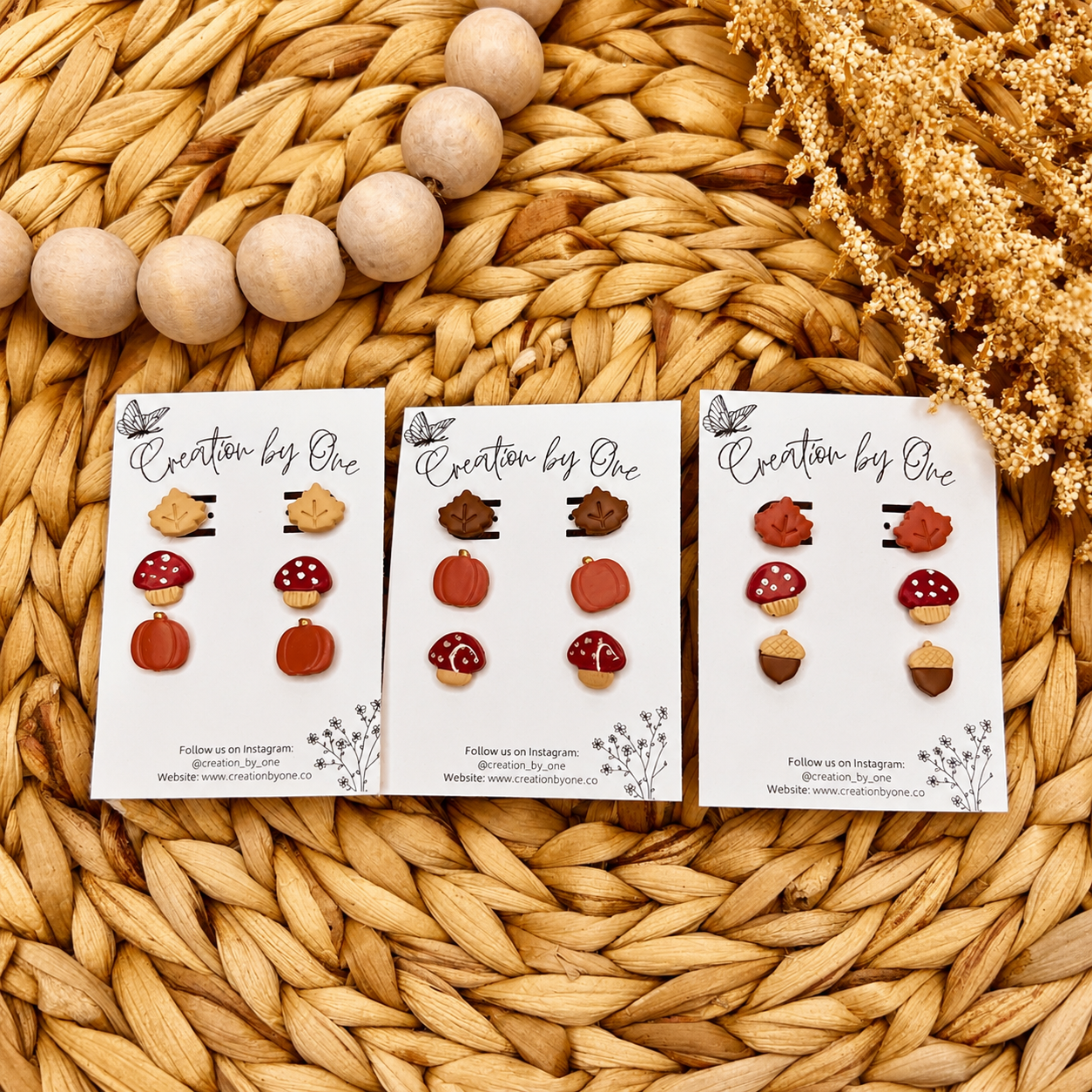 Handmade fall clay stud earrings by Creation by One featuring beige maple leaves, burgundy mushrooms, and dusty pumpkin studs displayed on a branded earring card with a neutral boho woven background, wooden beads, and dried florals.