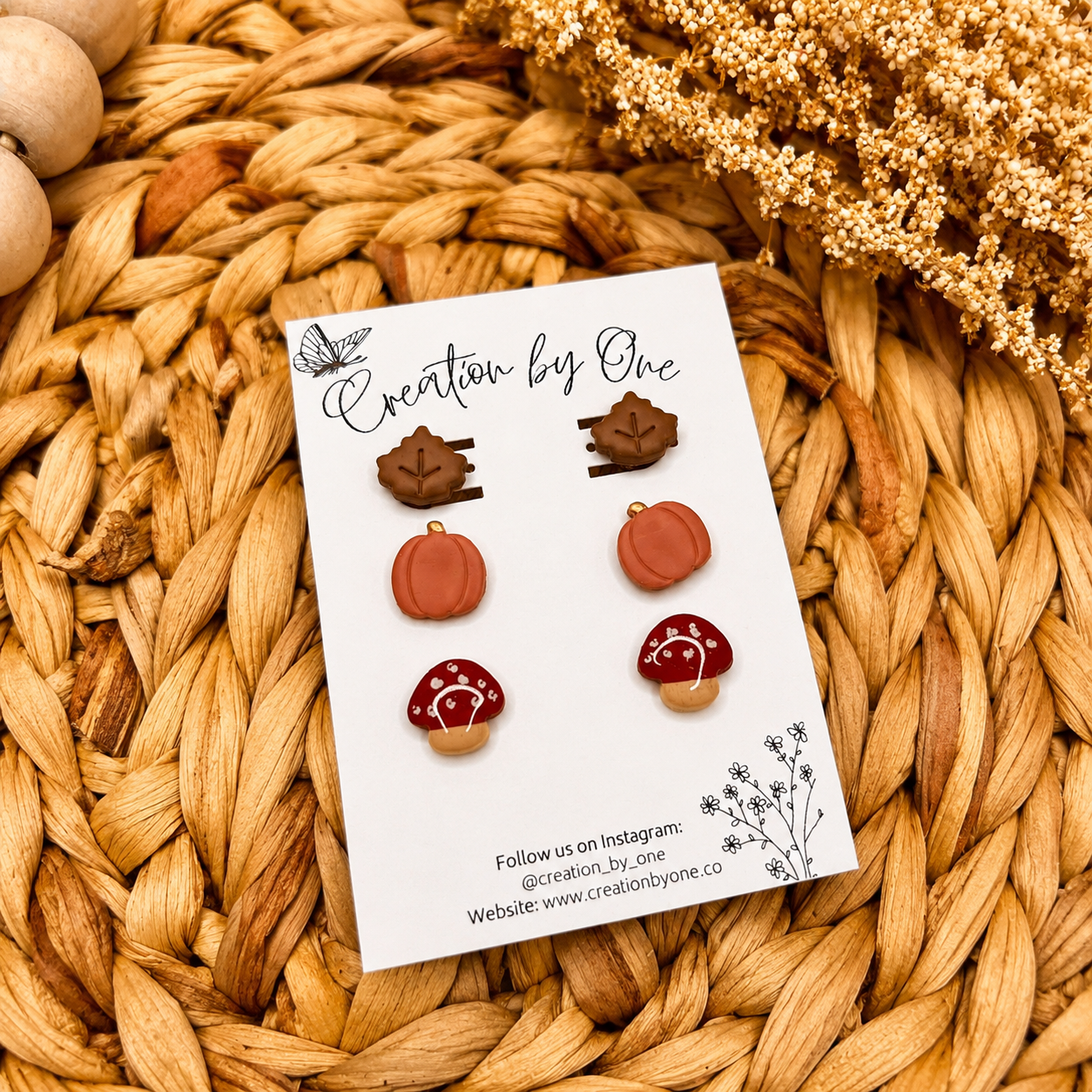 Handmade fall clay stud earrings by Creation by One featuring beige maple leaves, burgundy mushrooms, and dusty pumpkin studs displayed on a branded earring card with a neutral boho woven background, wooden beads, and dried florals.