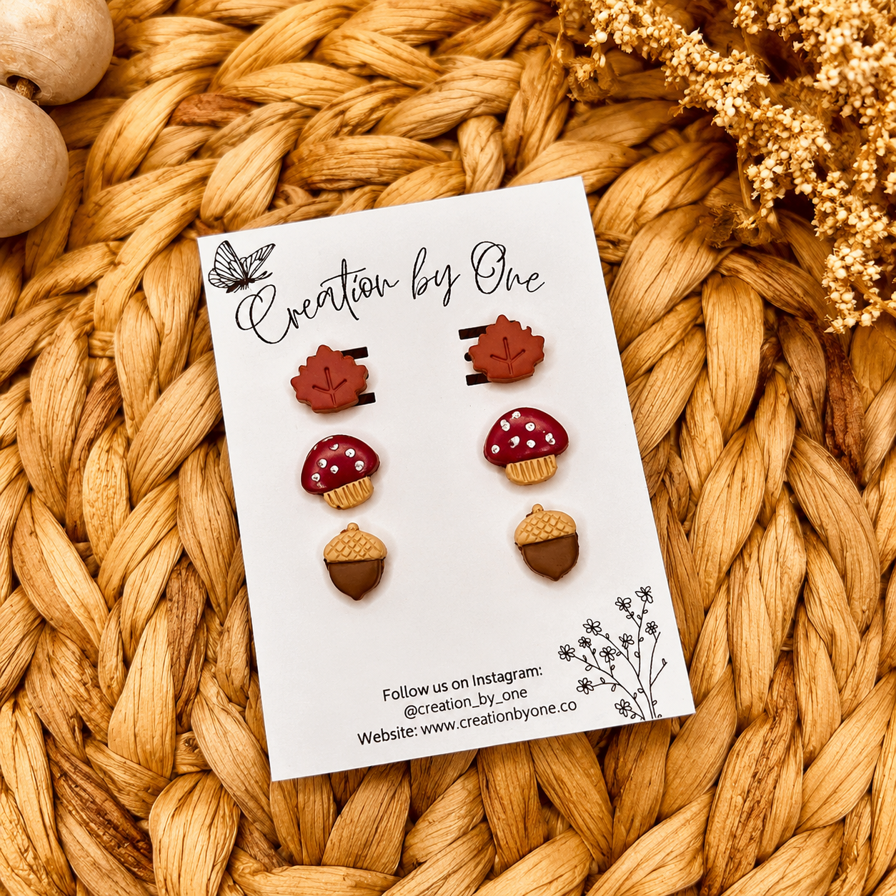 Handmade fall clay stud earrings by Creation by One featuring beige maple leaves, burgundy mushrooms, and dusty pumpkin studs displayed on a branded earring card with a neutral boho woven background, wooden beads, and dried florals.