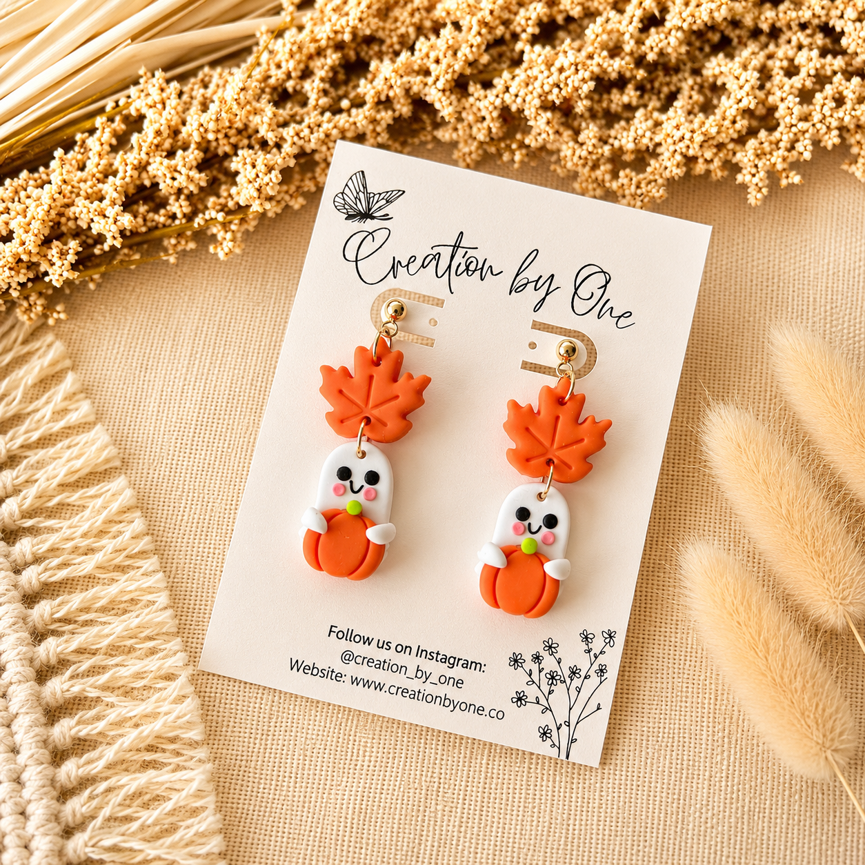 Pair of earrings on a 'Creation by One' branded card with dried plants in the background