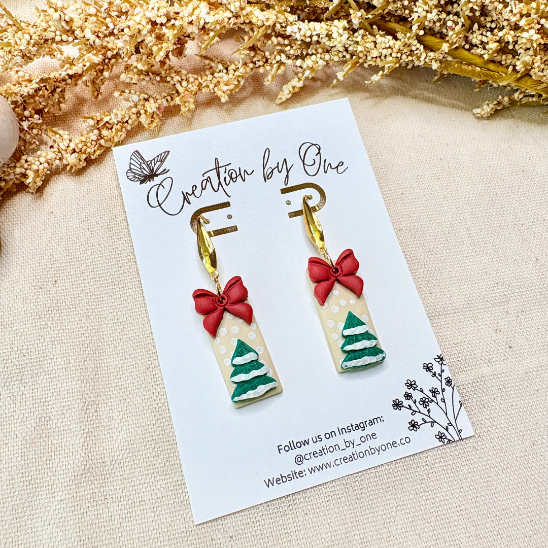 Festive polymer clay bow Christmas earrings displayed with soft snow and Christmas tree décor