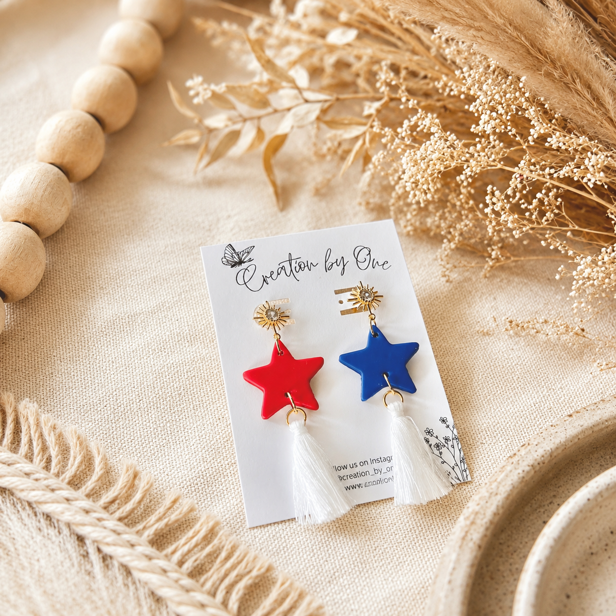 Pair of earrings with red and blue star designs on a branded card, surrounded by natural elements.
