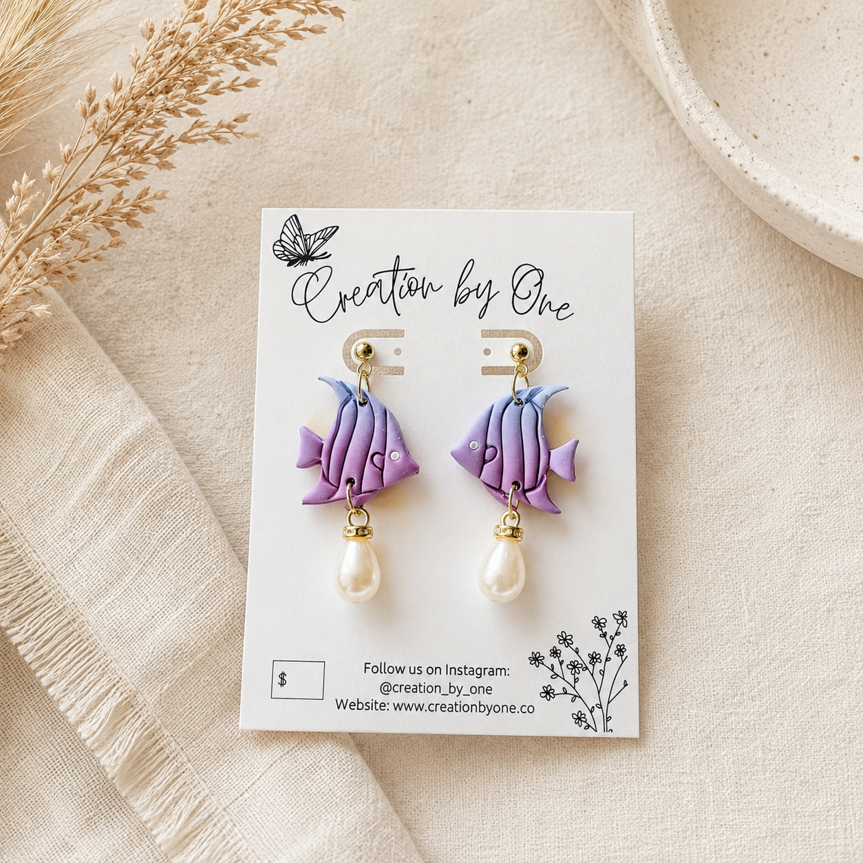 A pair of handmade polymer clay fish drop earrings in purple and blue, each with a white pearl dangle and gold post, displayed on a white card labeled 'Creation by One' on a beige linen background.