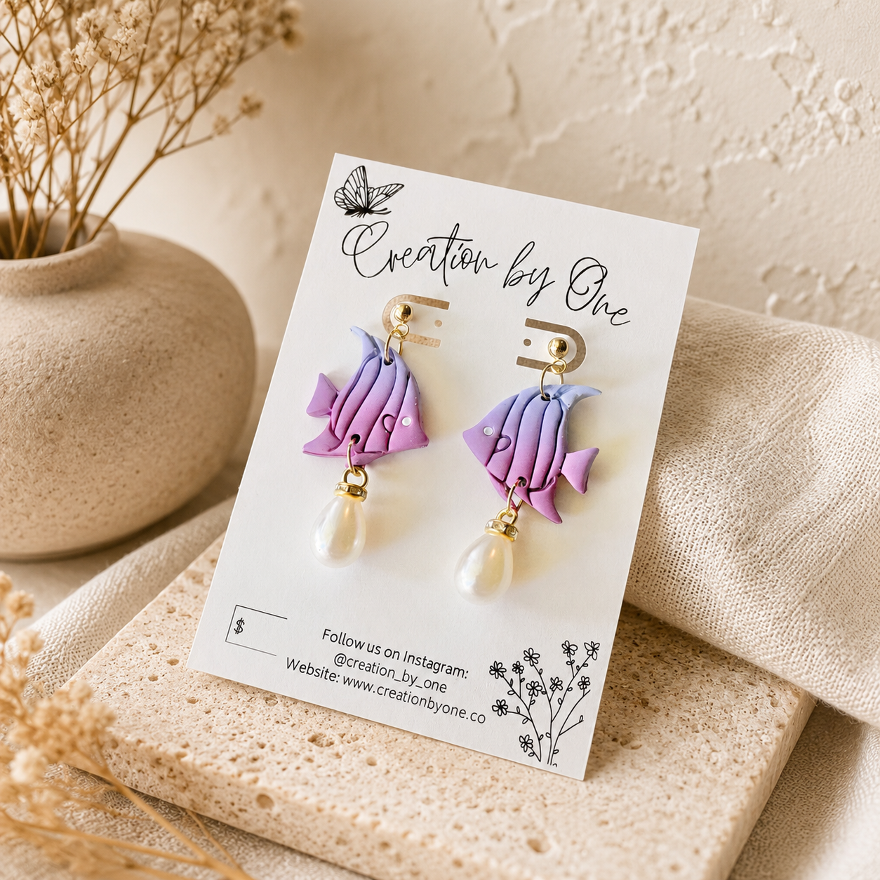 A pair of handmade polymer clay fish drop earrings in purple and lavender, displayed on a white card labeled 'Creation by One'. The earrings feature gold-plated hooks and dangling faux pearls, set against a styled background of beige linen, a ceramic vase with dried flowers, and textured stone.