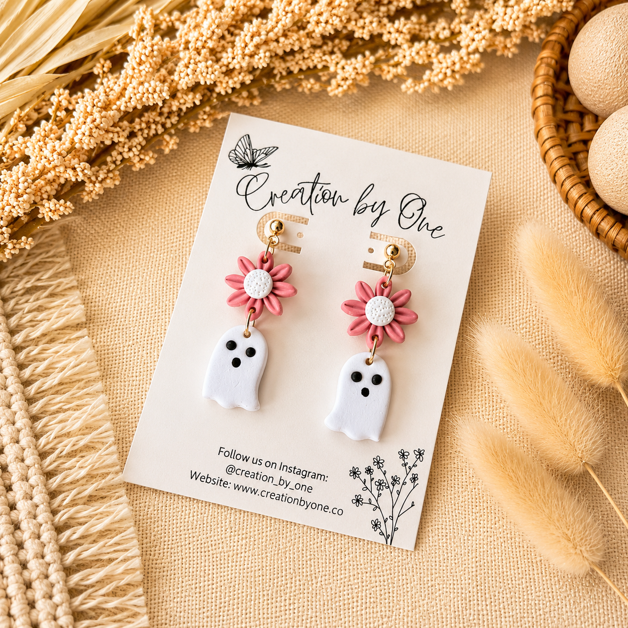 Pair of ghost-shaped earrings with pink flower details on a 'Creation by One' branded card, surrounded by dried plants.