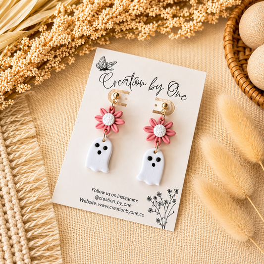 Pair of ghost-shaped earrings with pink flower details on a 'Creation by One' branded card, surrounded by dried plants.
