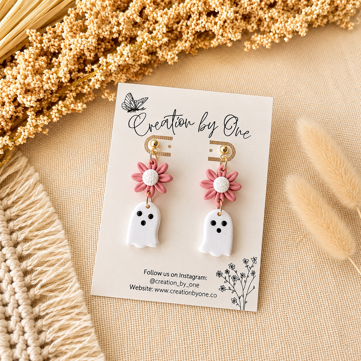 Pair of earrings on a 'Creation by One' branded card with dried flowers in the background.