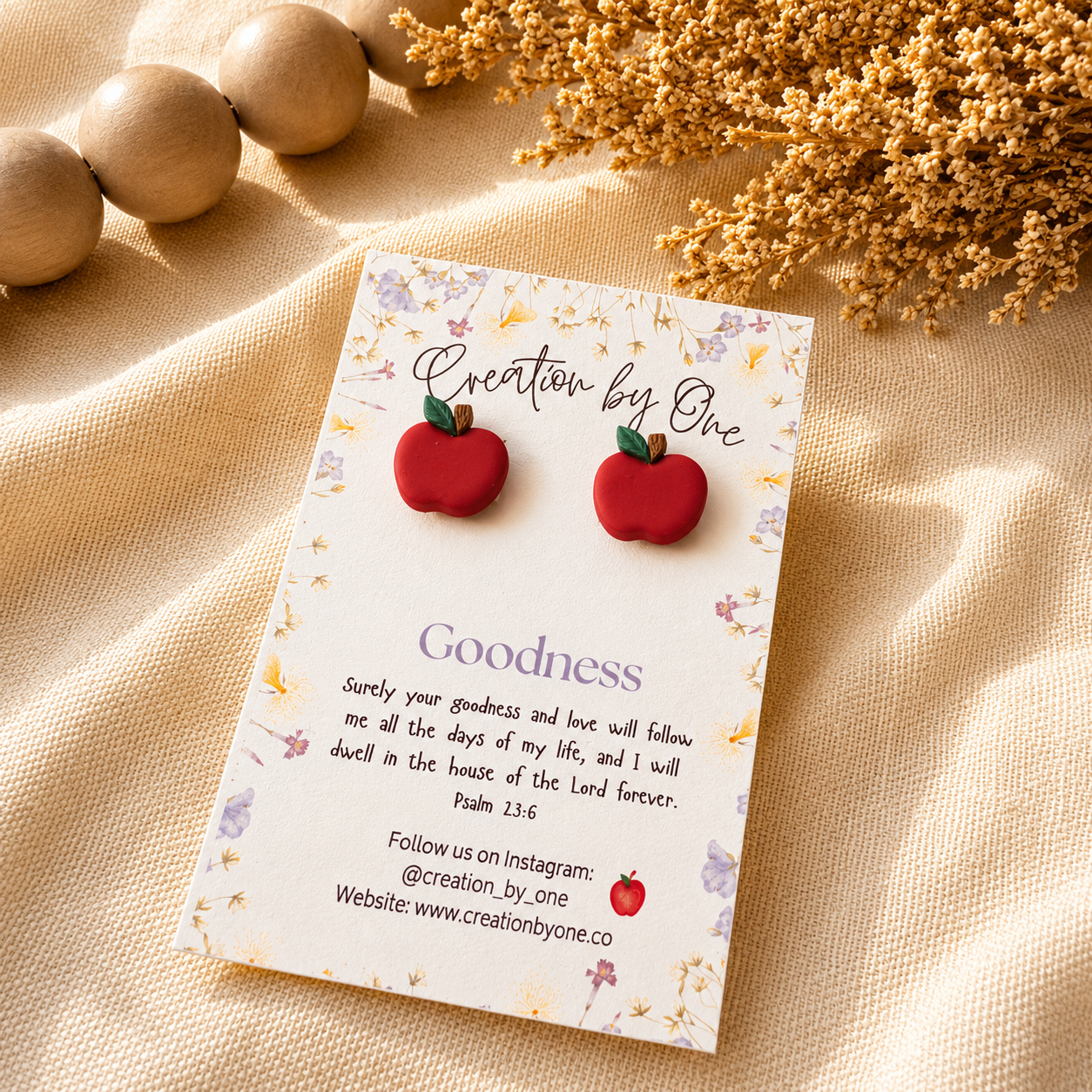 Red apple-shaped earrings on a 'Creation by One' branded card with decorative elements on a beige fabric background.