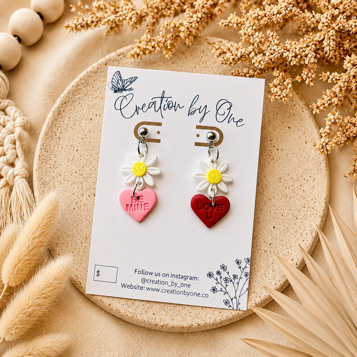Handmade daisy heart dangle earrings featuring white flower charms with pink “Be Mine” and red “Love U” hearts, displayed on a Creation by One jewelry card against a soft neutral linen background with dried florals in a modern boho flat lay.