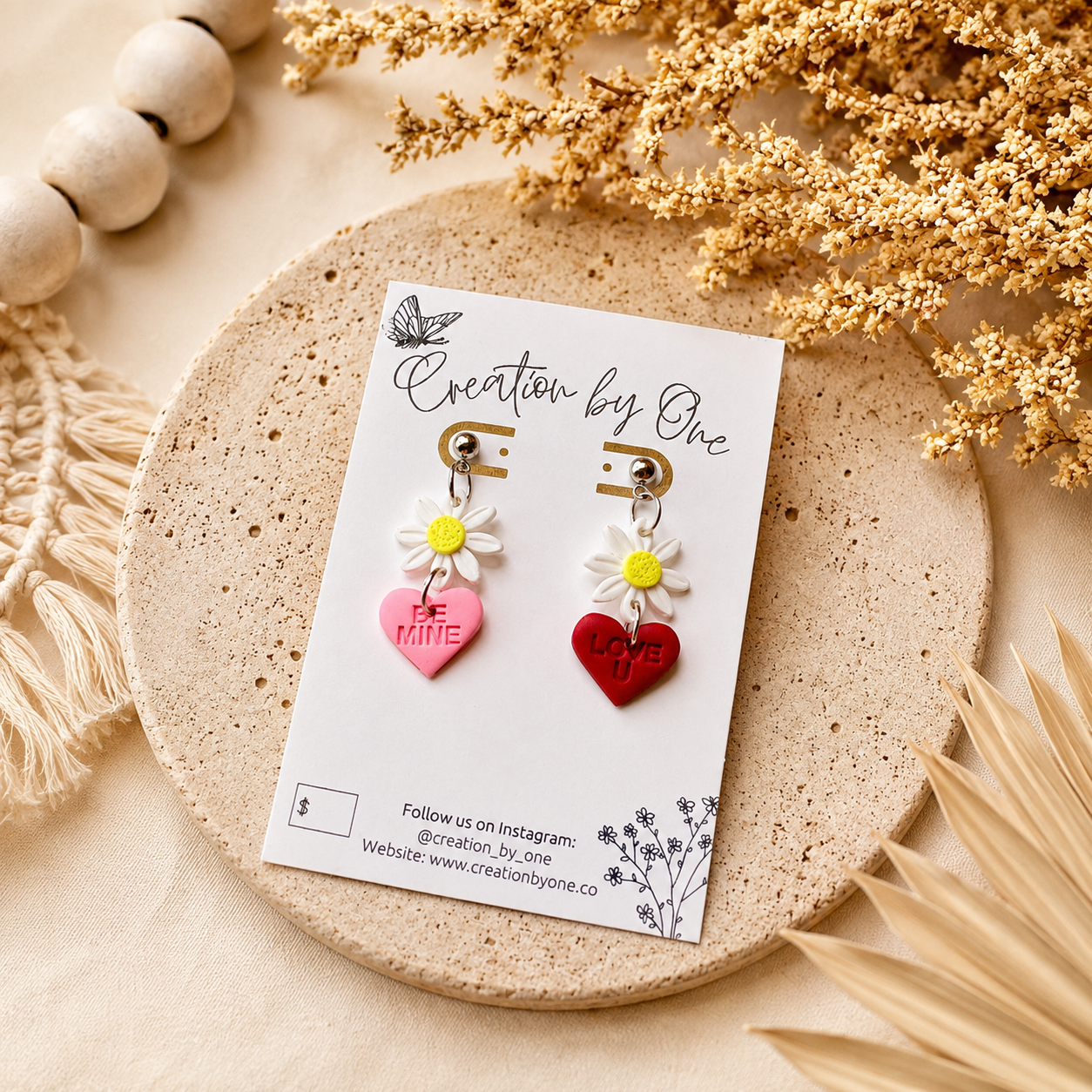 Handmade daisy heart dangle earrings featuring white flower charms with pink “Be Mine” and red “Love U” hearts, displayed on a Creation by One jewelry card against a soft neutral linen background with dried florals in a modern boho flat lay.