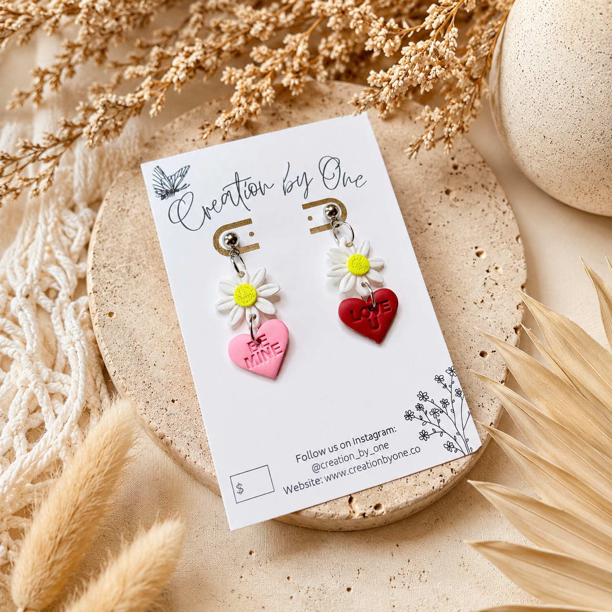 Handmade daisy heart dangle earrings featuring white flower charms with pink “Be Mine” and red “Love U” hearts, displayed on a Creation by One jewelry card against a soft neutral linen background with dried florals in a modern boho flat lay.