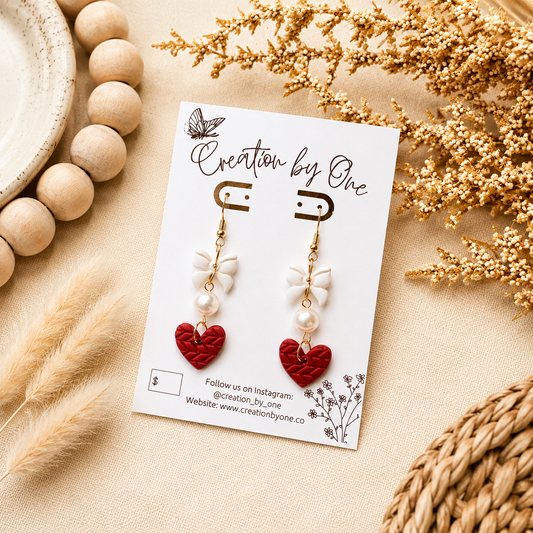 Handmade red heart dangle earrings with white bows and faux pearl accents displayed on a jewelry card against a soft neutral linen background with dried florals. Boho modern flat lay product photography featuring lightweight Valentine’s Day statement earrings with gold hooks, perfect for gifting.