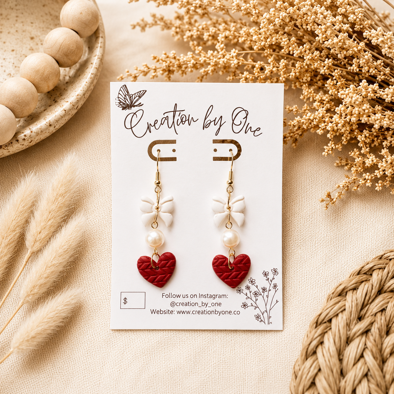 Handmade red heart dangle earrings with white bows and faux pearl accents displayed on a jewelry card against a soft neutral linen background with dried florals. Boho modern flat lay product photography featuring lightweight Valentine’s Day statement earrings with gold hooks, perfect for gifting.