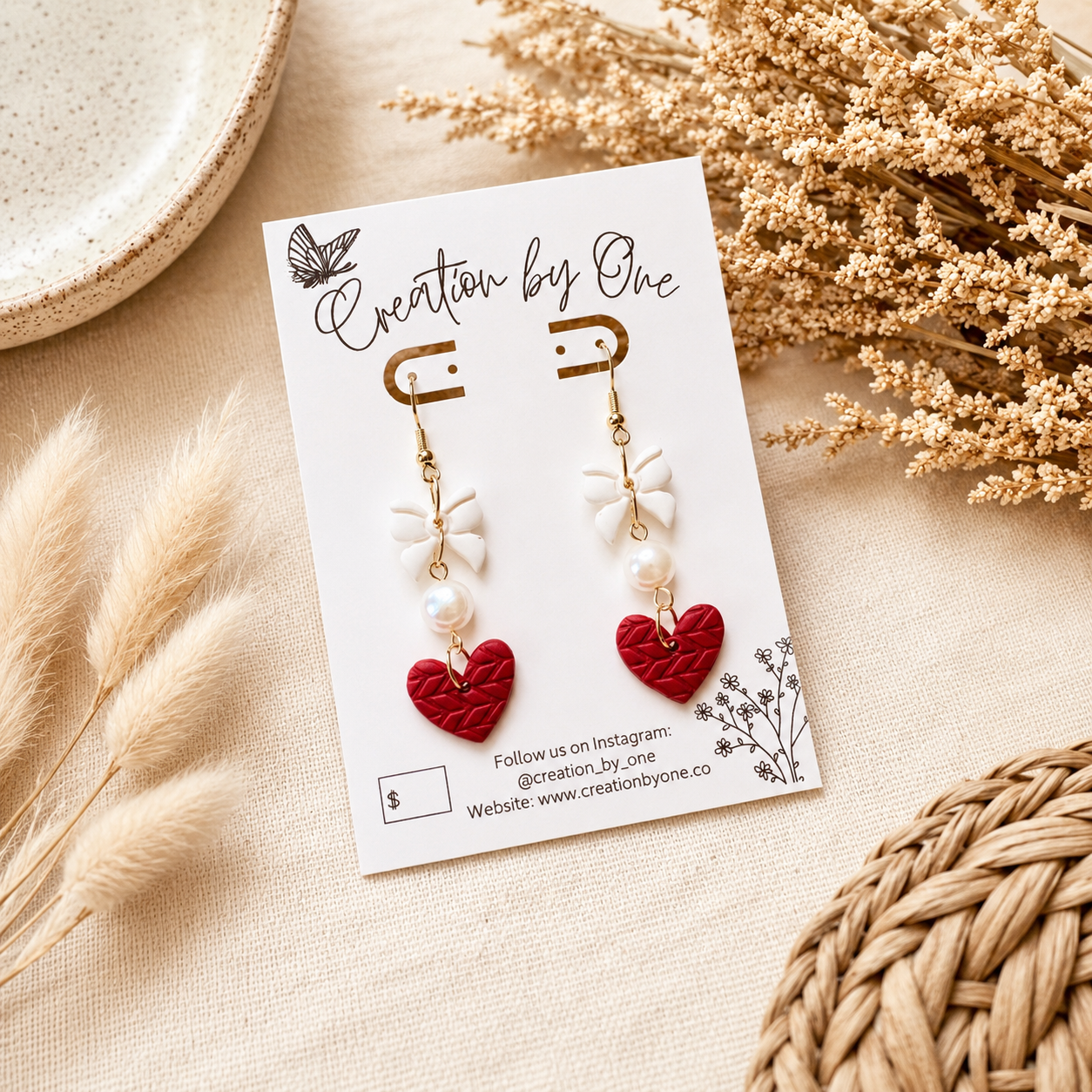 Handmade red heart dangle earrings with white bows and faux pearl accents displayed on a jewelry card against a soft neutral linen background with dried florals. Boho modern flat lay product photography featuring lightweight Valentine’s Day statement earrings with gold hooks, perfect for gifting.