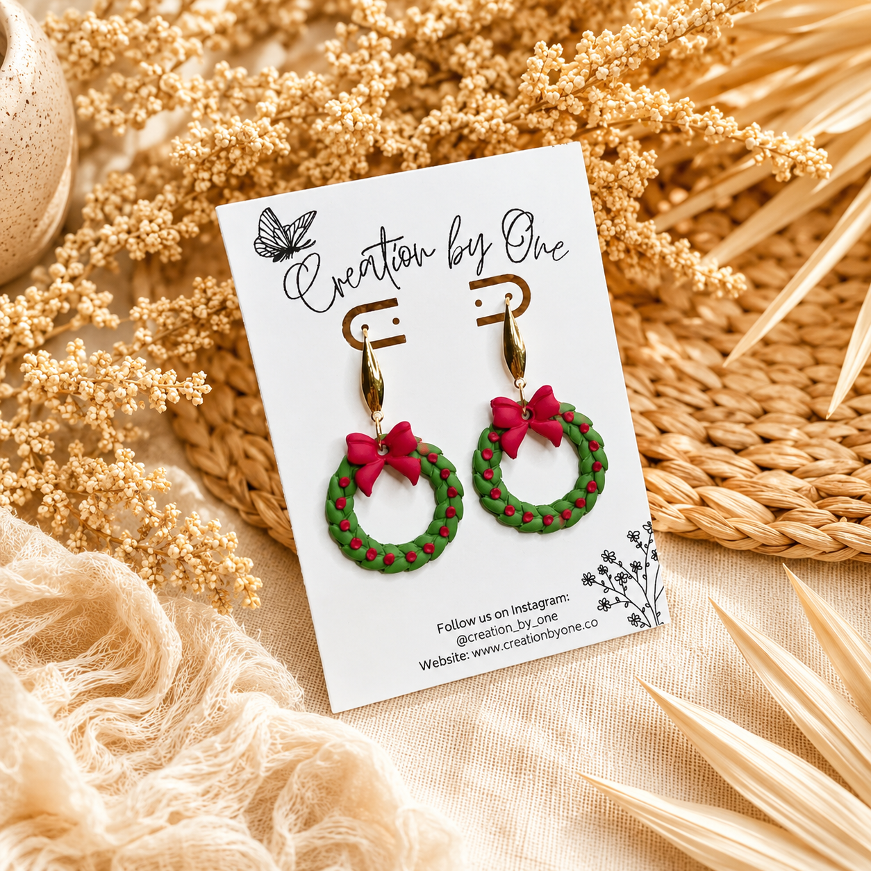 Handmade Christmas wreath dangle earrings with gold teardrop hooks, featuring green wreaths with red berry accents and bright red bows. Displayed on a Creation by One jewelry card with a warm boho background of dried florals and neutral linen, perfect for festive holiday outfits and Christmas gift giving.