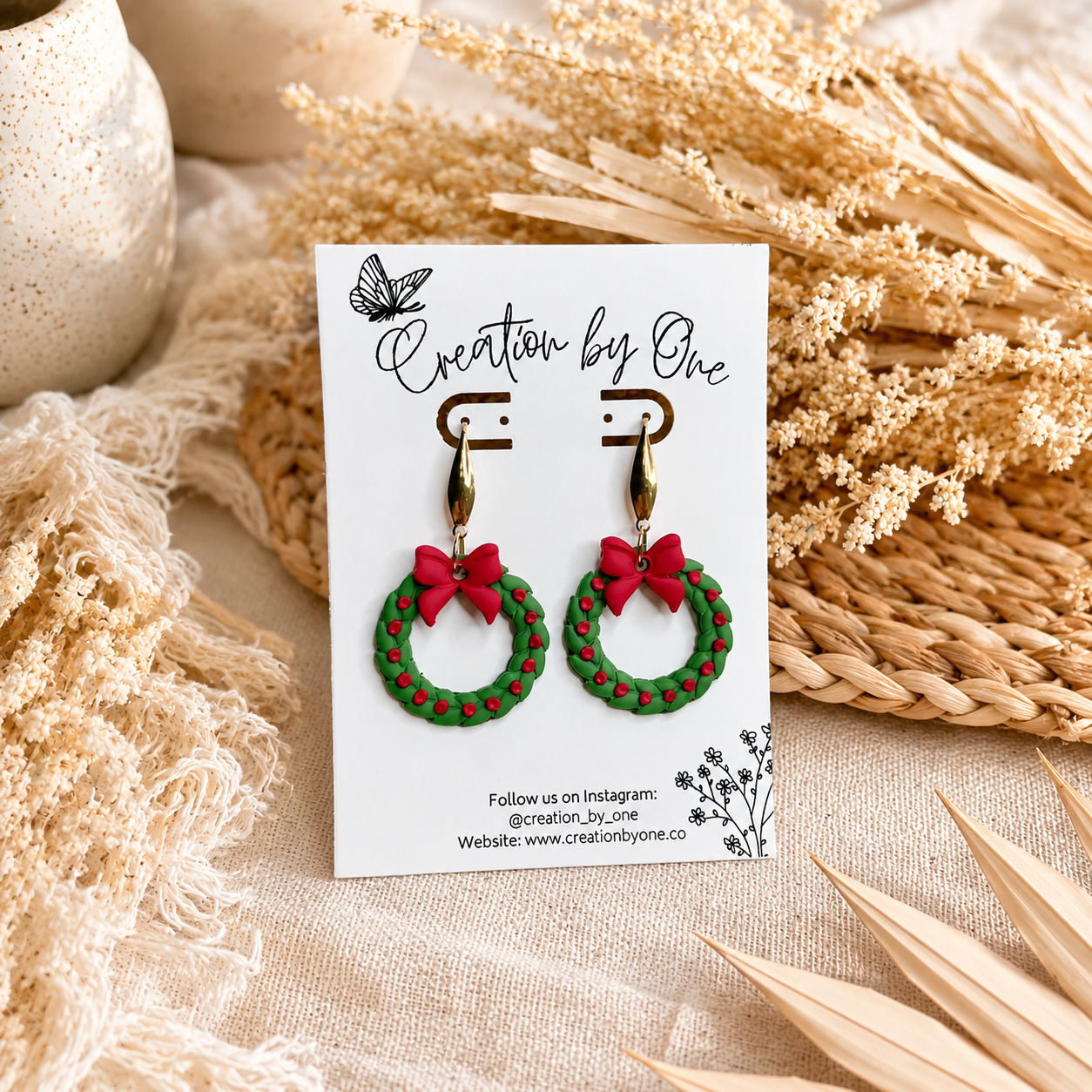 Handmade Christmas wreath dangle earrings with gold teardrop hooks, featuring green wreaths with red berry accents and bright red bows. Displayed on a Creation by One jewelry card with a warm boho background of dried florals and neutral linen, perfect for festive holiday outfits and Christmas gift giving.