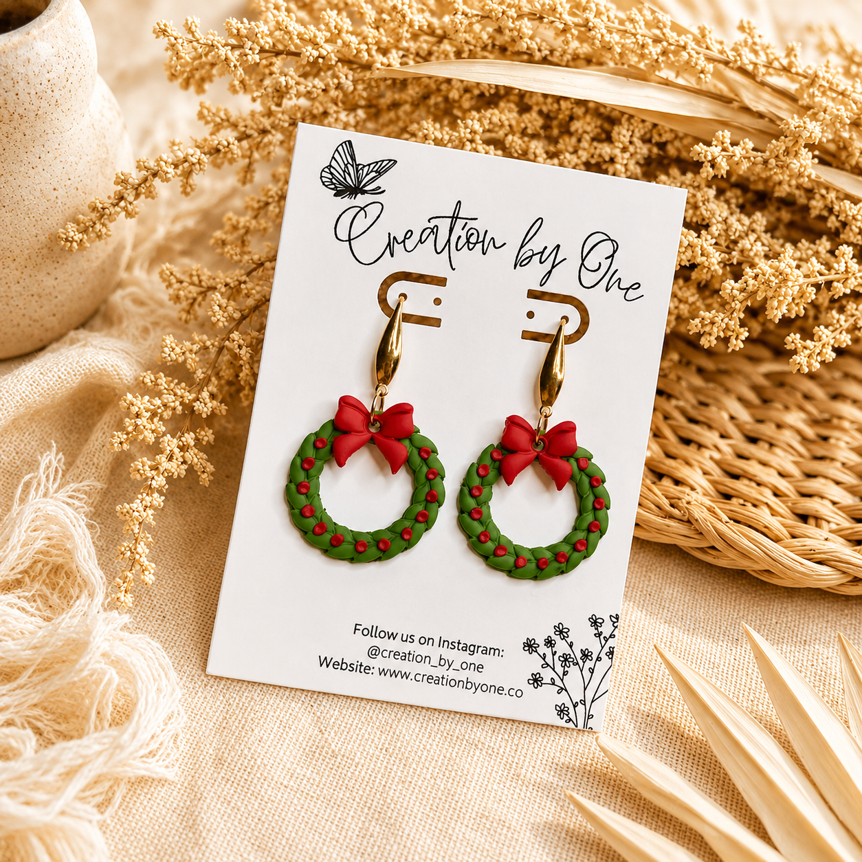 Handmade Christmas wreath dangle earrings with gold teardrop hooks, featuring green wreaths with red berry accents and bright red bows. Displayed on a Creation by One jewelry card with a warm boho background of dried florals and neutral linen, perfect for festive holiday outfits and Christmas gift giving.