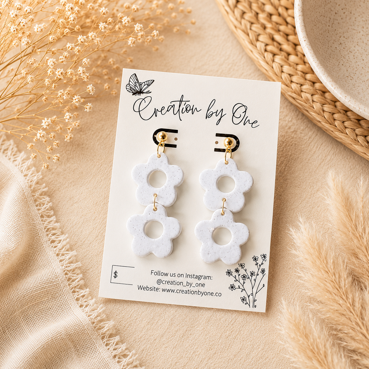 A pair of white flower-shaped drop earrings with gold-tone posts, displayed on a white card labeled 'Creation by One' and styled with dried flowers and a woven mat.