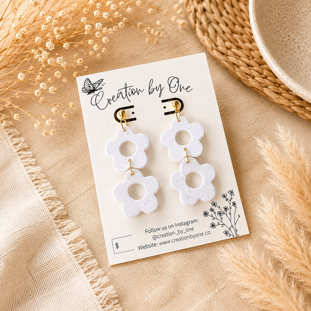 A pair of white flower-shaped drop earrings with gold-tone hooks, displayed on a branded card against a neutral beige fabric background with dried floral props.