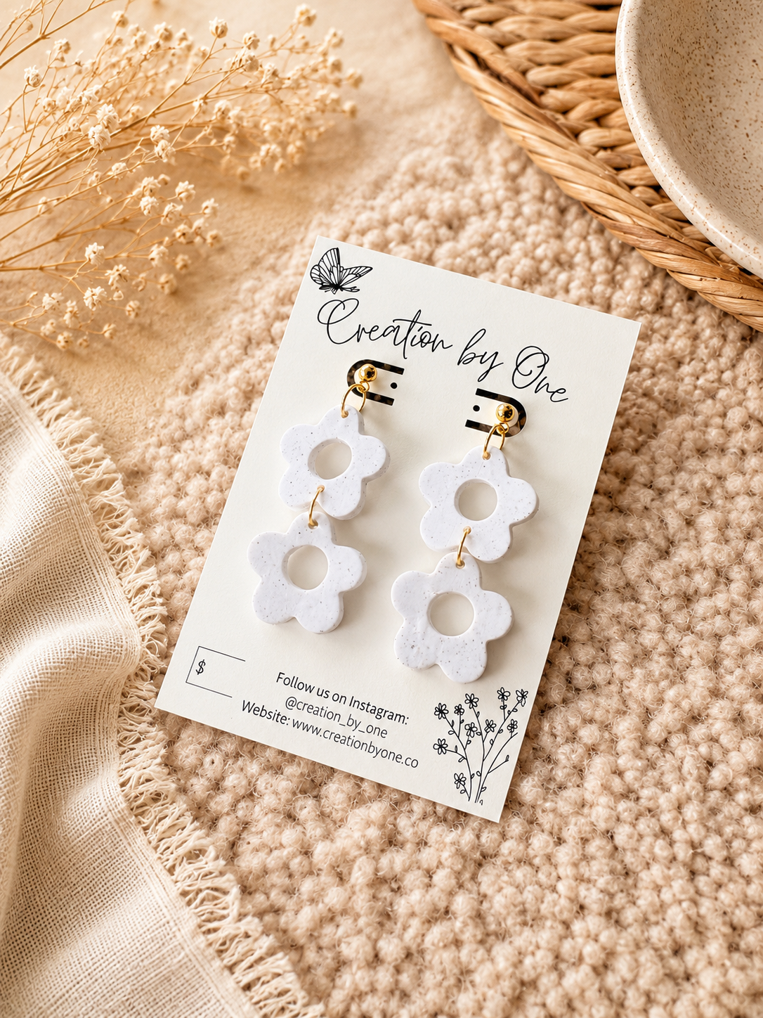 White flower-shaped drop earrings with gold-tone posts, displayed on a branded card over a beige textured blanket with dried flowers and woven decor.