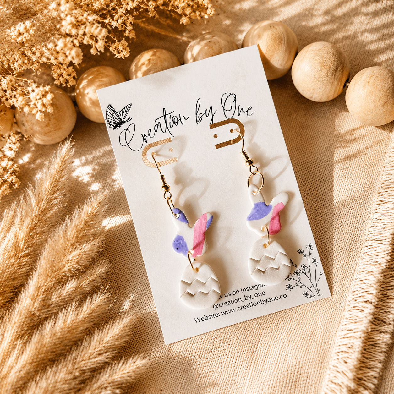 Handmade Easter bunny and egg polymer clay dangle earrings by Creation by One displayed on a neutral boho fabric background with natural wooden beads, dried florals, and warm sunlight. Lightweight spring earrings in pastel pink, lavender, and white, perfect for Easter baskets, church, and seasonal outfits.