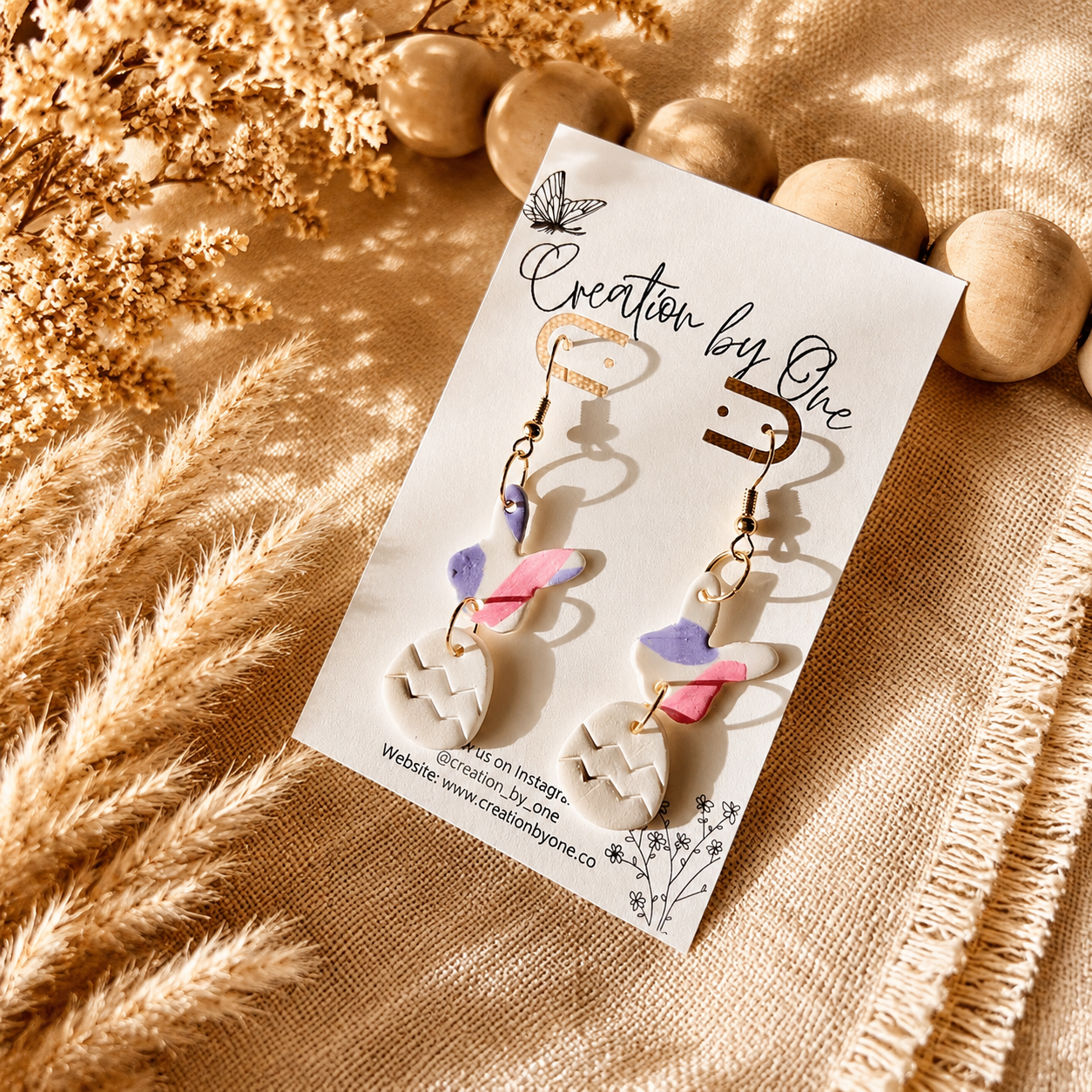 Handmade Easter bunny and egg polymer clay dangle earrings by Creation by One displayed on a neutral boho fabric background with natural wooden beads, dried florals, and warm sunlight. Lightweight spring earrings in pastel pink, lavender, and white, perfect for Easter baskets, church, and seasonal outfits.