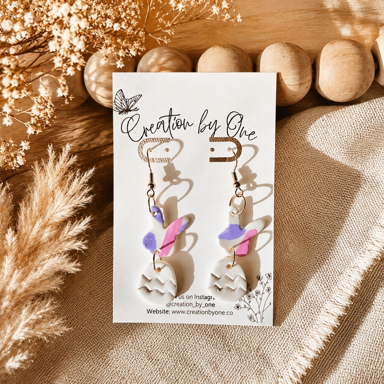 Handmade Easter bunny and egg polymer clay dangle earrings by Creation by One displayed on a neutral boho fabric background with natural wooden beads, dried florals, and warm sunlight. Lightweight spring earrings in pastel pink, lavender, and white, perfect for Easter baskets, church, and seasonal outfits.