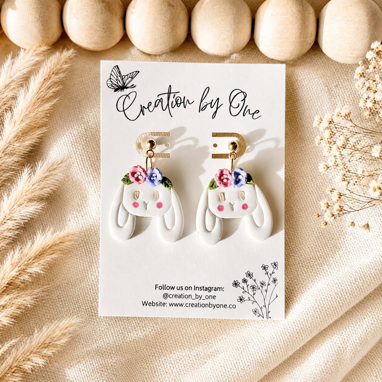 Handmade white bunny dangle earrings with pink and purple floral crowns on gold posts displayed on a Creation by One jewelry card against a neutral boho linen background with wooden beads, perfect for Easter, spring outfits, and cottagecore fashion.