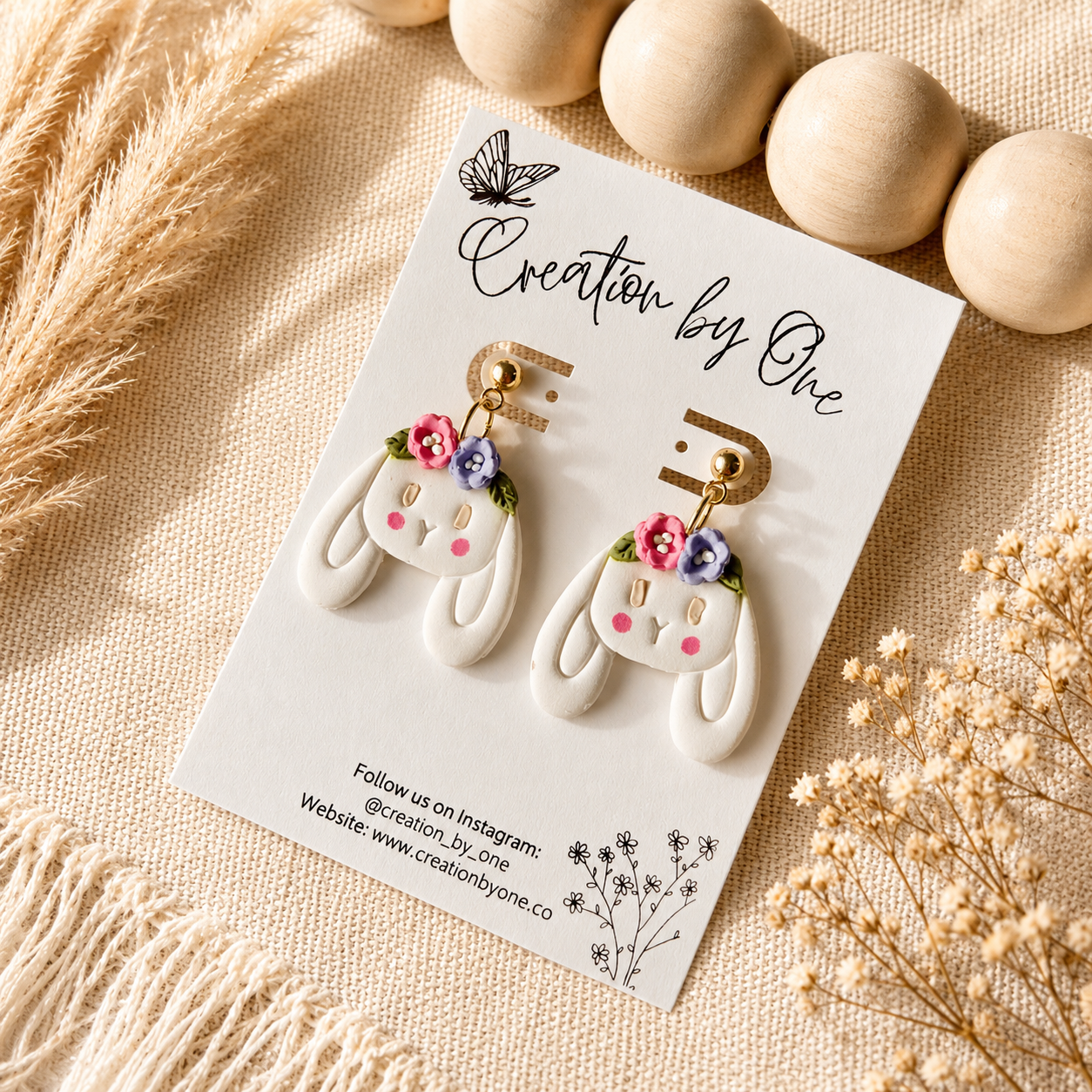 Handmade white bunny dangle earrings with pink and purple floral crowns on gold posts displayed on a Creation by One jewelry card against a neutral boho linen background with wooden beads, perfect for Easter, spring outfits, and cottagecore fashion.