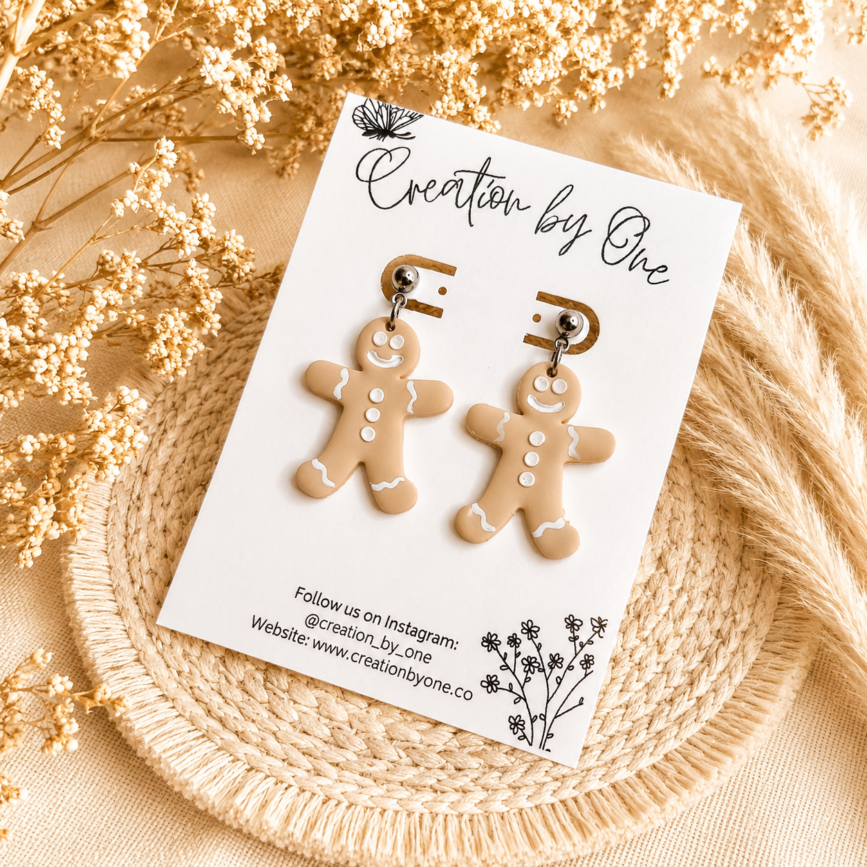 Handmade gingerbread man dangle earrings on stainless steel studs displayed on a Creation by One jewelry card with a neutral boho background featuring dried florals and woven natural textures. Festive Christmas clay earrings perfect for holiday outfits, stocking stuffers, and seasonal gifts.