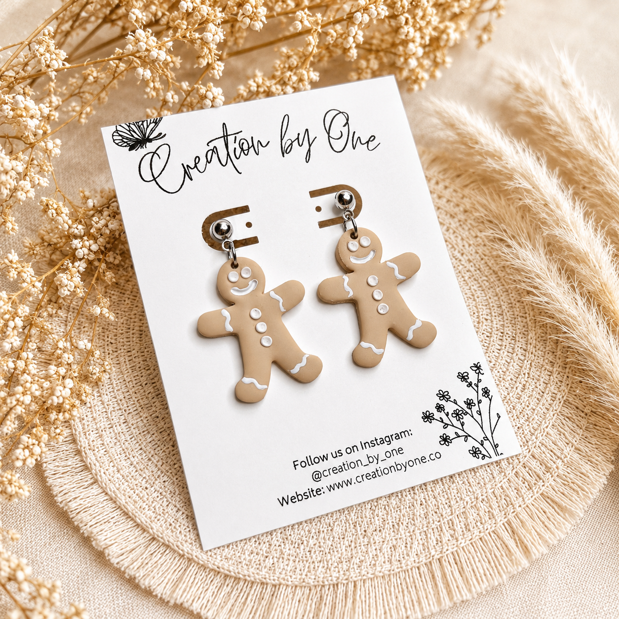 Handmade gingerbread man dangle earrings on stainless steel studs displayed on a Creation by One jewelry card with a neutral boho background featuring dried florals and woven natural textures. Festive Christmas clay earrings perfect for holiday outfits, stocking stuffers, and seasonal gifts.