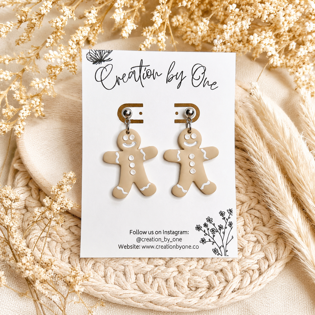 Handmade gingerbread man dangle earrings on stainless steel studs displayed on a Creation by One jewelry card with a neutral boho background featuring dried florals and woven natural textures. Festive Christmas clay earrings perfect for holiday outfits, stocking stuffers, and seasonal gifts.