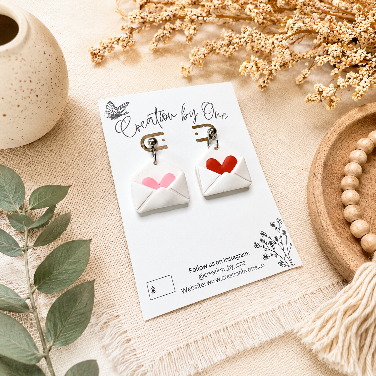 Handmade envelope heart dangle earrings displayed on a Creation by One jewelry card, styled on a neutral linen background with dried florals and natural wood beads in a modern boho flat lay. Perfect Valentine’s Day gift jewelry.