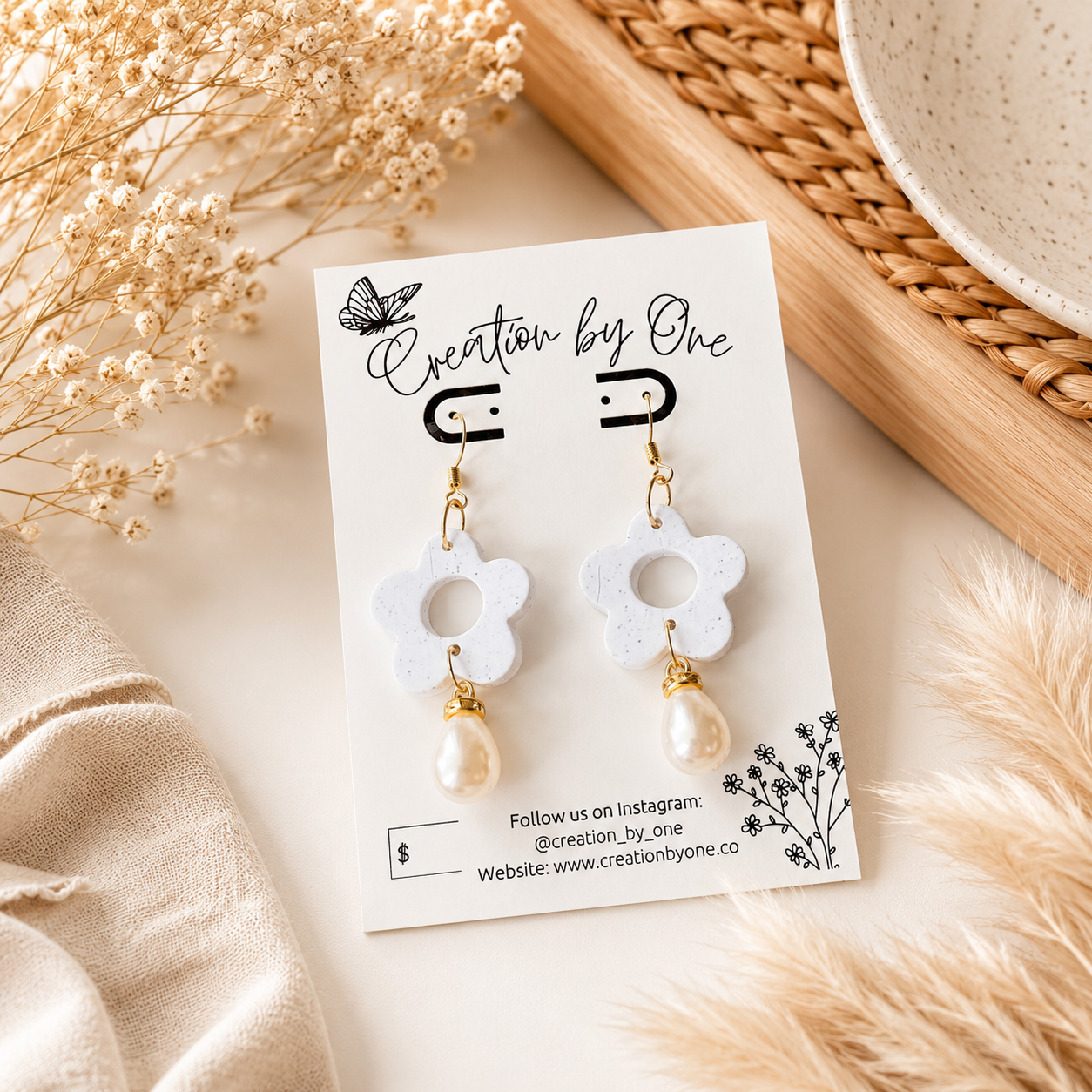 A pair of white flower-shaped drop earrings with pearl-like dangling beads and gold-tone hooks, displayed on a branded card. The card is styled on a light surface with dried flowers, fabric, and a woven tray in the background.