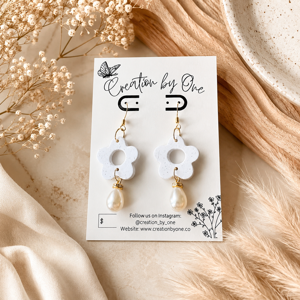 A pair of white flower-shaped drop earrings with gold-tone hooks and pearl-like dangling beads, displayed on a branded card. The card is styled in a warm, neutral flat-lay setting with dried flowers, beige fabric, and a ceramic dish.
