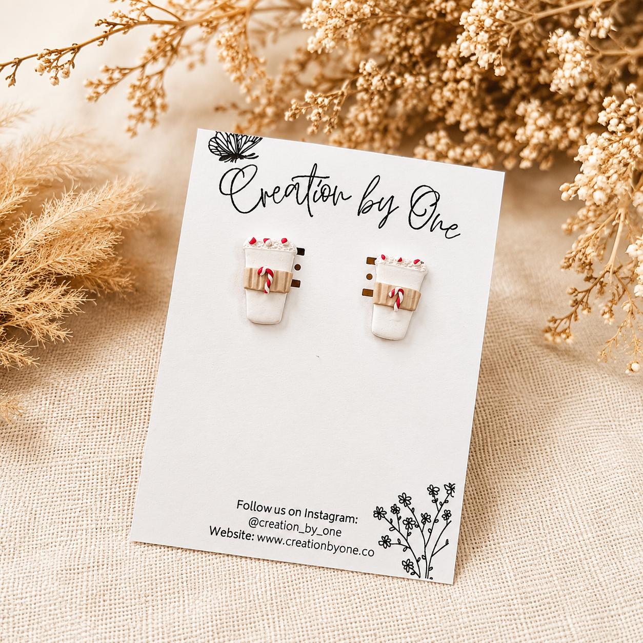 Handmade white polymer clay coffee cup stud earrings with peppermint candy cane details displayed on a branded jewelry card, styled on a neutral linen background with dried boho florals for a cozy Christmas aesthetic.