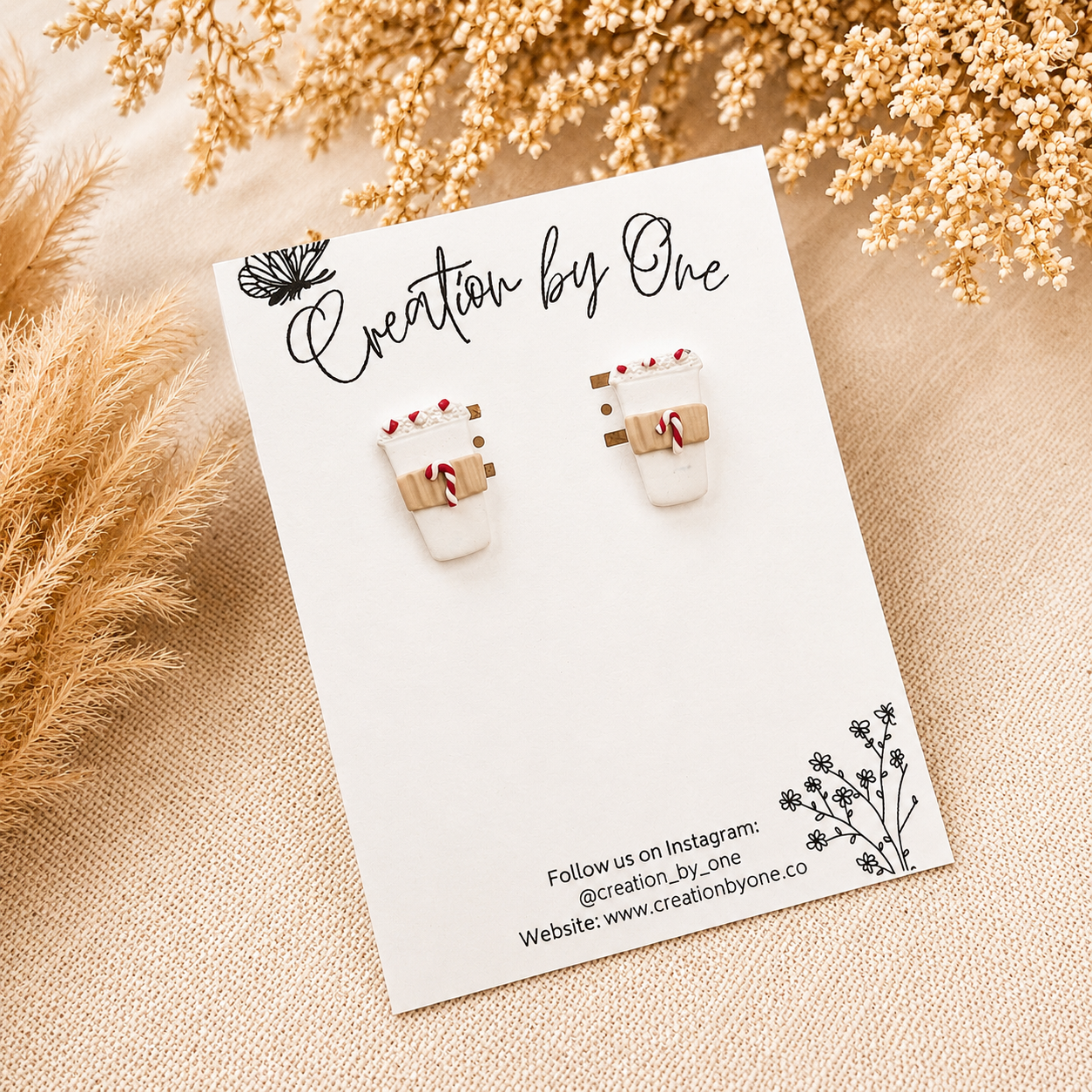 Handmade white polymer clay coffee cup stud earrings with peppermint candy cane details displayed on a branded jewelry card, styled on a neutral linen background with dried boho florals for a cozy Christmas aesthetic.