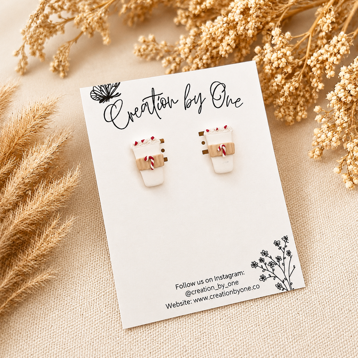 Handmade white polymer clay coffee cup stud earrings with peppermint candy cane details displayed on a branded jewelry card, styled on a neutral linen background with dried boho florals for a cozy Christmas aesthetic.