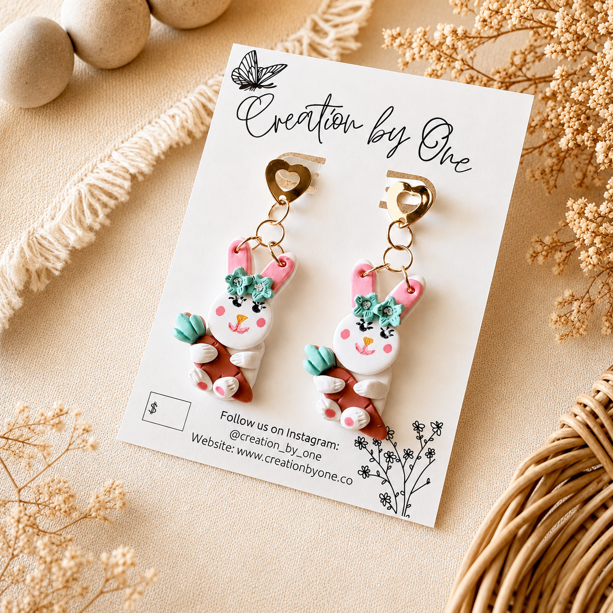 Handmade pink bunny carrot dangle earrings with gold heart studs on a Creation by One display card, styled on a neutral boho linen background with dried florals for Easter spring jewelry photography.