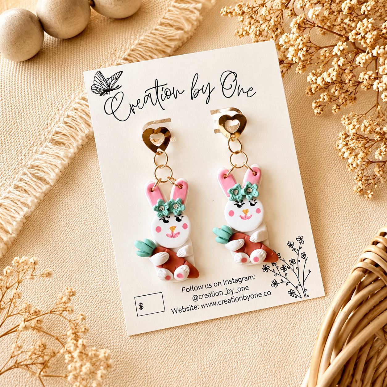 Handmade pink bunny carrot dangle earrings with gold heart studs on a Creation by One display card, styled on a neutral boho linen background with dried florals for Easter spring jewelry photography.