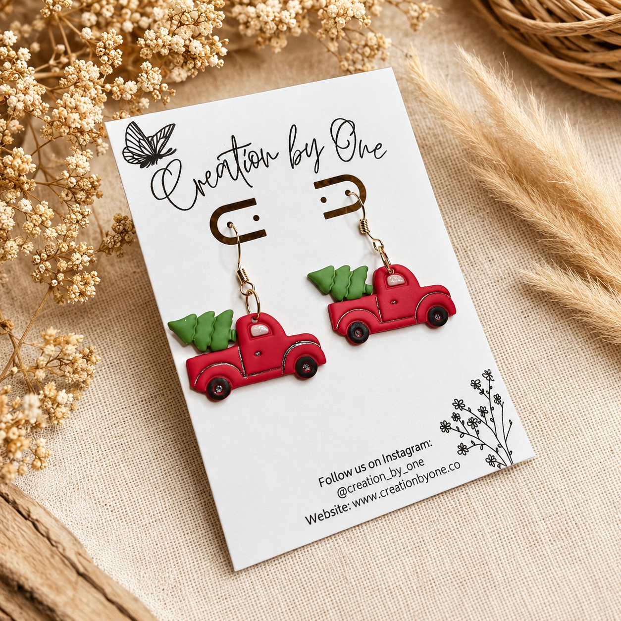 Handmade red vintage truck Christmas tree dangle earrings on gold hooks displayed on a Creation by One card with a neutral boho linen background, dried florals, and rustic farmhouse decor.