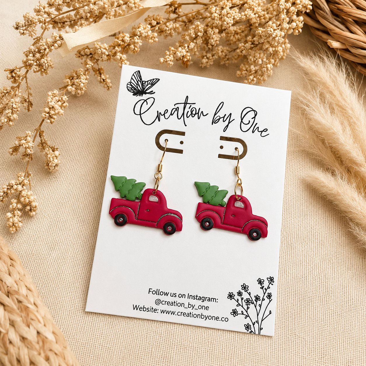 Handmade red vintage truck Christmas tree dangle earrings on gold hooks displayed on a Creation by One card with a neutral boho linen background, dried florals, and rustic farmhouse decor.