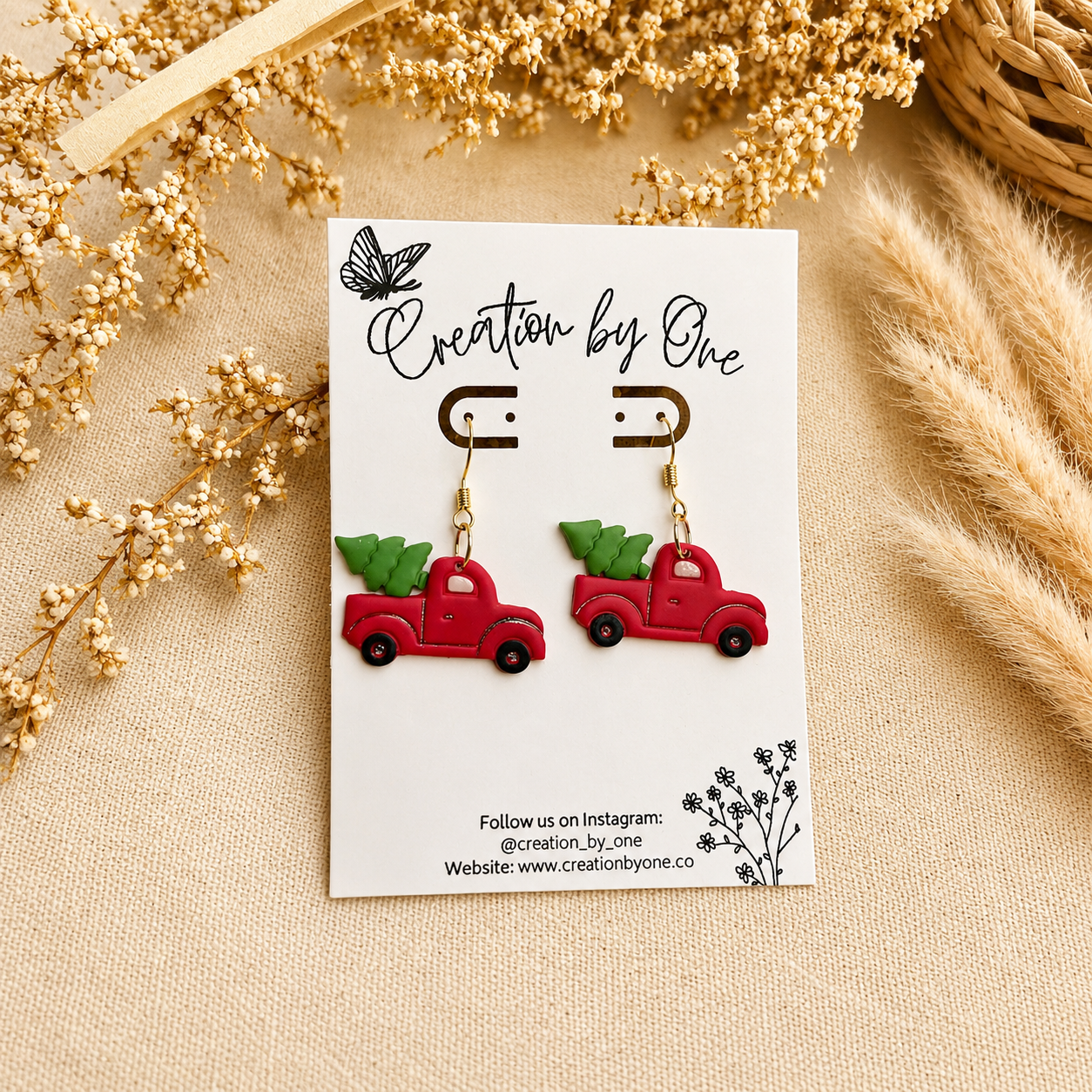 Handmade red vintage truck Christmas tree dangle earrings on gold hooks displayed on a Creation by One card with a neutral boho linen background, dried florals, and rustic farmhouse decor.
