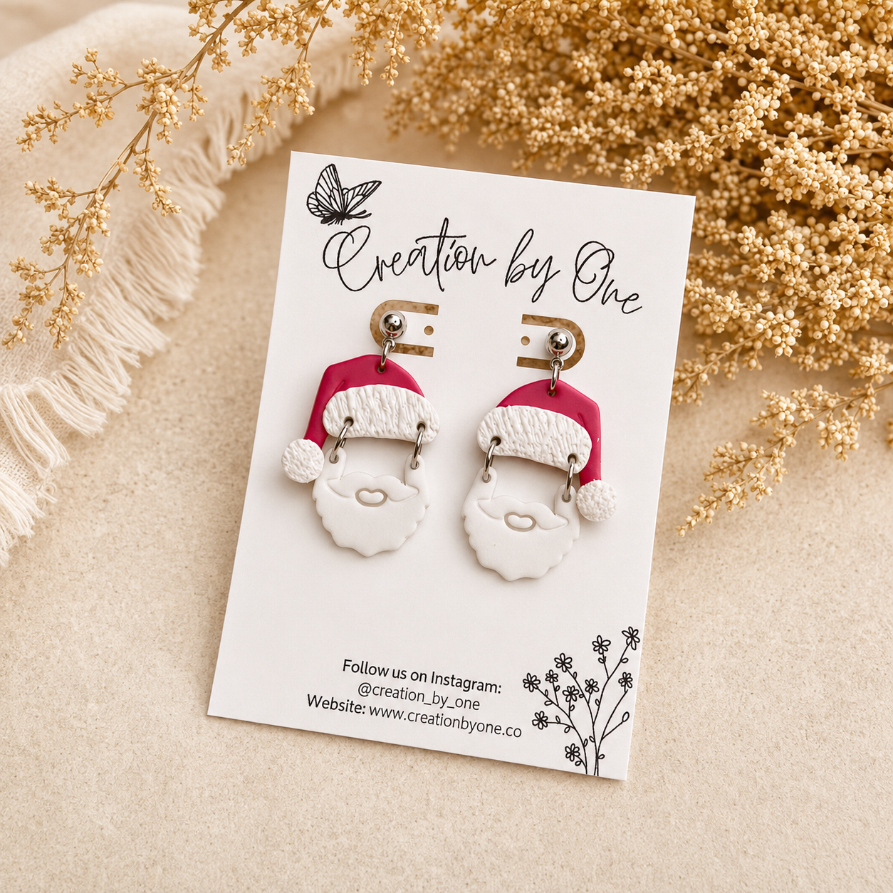 Handmade Santa Claus polymer clay dangle earrings on a Creation by One display card with a neutral boho background featuring dried florals and soft beige textures, perfect for Christmas outfits, holiday gifting, and festive winter accessories.