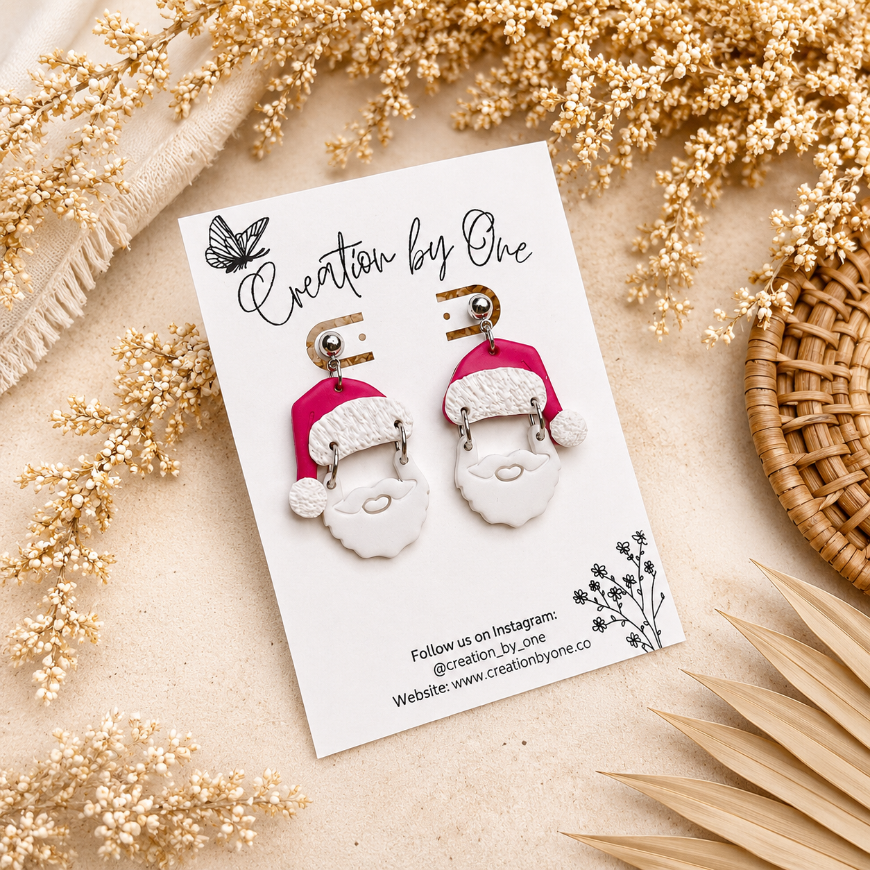 Handmade Santa Claus polymer clay dangle earrings on a Creation by One display card with a neutral boho background featuring dried florals and soft beige textures, perfect for Christmas outfits, holiday gifting, and festive winter accessories.